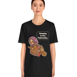 Puede incluir: Una camiseta negra de manga corta con una ilustraci&oacute;n gr&aacute;fica de una persona mayor con un pa&ntilde;uelo morado y gafas, montada en una escoba. El gr&aacute;fico incluye un bocadillo con el texto: Sweeping in late. Fashionably.