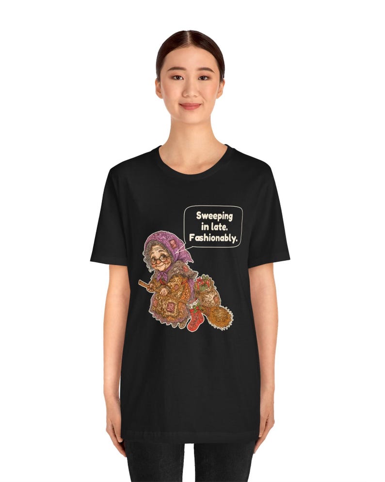 Puede incluir: Una camiseta negra de manga corta con una ilustraci&oacute;n gr&aacute;fica de una persona mayor con un pa&ntilde;uelo morado y gafas, montada en una escoba. El gr&aacute;fico incluye un bocadillo con el texto: Sweeping in late. Fashionably.