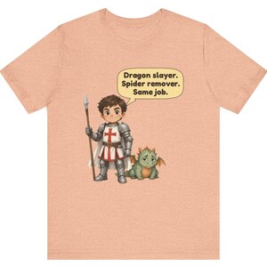 K&ouml;nnte beinhalten: Ein pfirsichfarbenes T-Shirt mit einem Cartoon-Ritter in silberner R&uuml;stung, der einen Speer h&auml;lt und neben einem kleinen gr&uuml;nen Drachen steht. Eine Sprechblase &uuml;ber dem Ritter enth&auml;lt den Text: Dragon slayer. Spider remover. Same job.