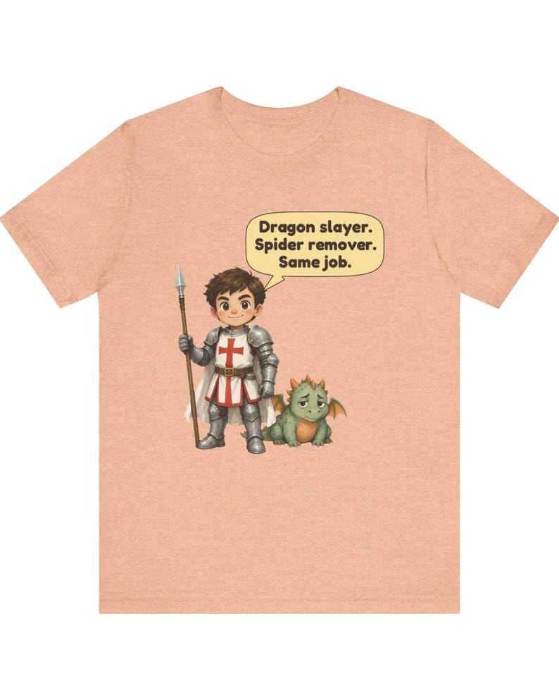 K&ouml;nnte beinhalten: Ein pfirsichfarbenes T-Shirt mit einem Cartoon-Ritter in silberner R&uuml;stung, der einen Speer h&auml;lt und neben einem kleinen gr&uuml;nen Drachen steht. Eine Sprechblase &uuml;ber dem Ritter enth&auml;lt den Text: Dragon slayer. Spider remover. Same job.