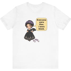 May include: A white t-shirt featuring a cartoon illustration of a character in a black dress with a lilac flower crown and a black fan. A speech bubble contains the text: Everyone wore lilac. I wore black. A lilac ribbon lies below.