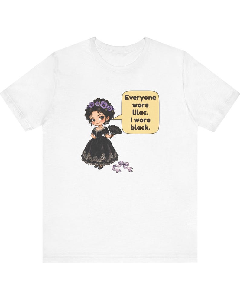 May include: A white t-shirt featuring a cartoon illustration of a character in a black dress with a lilac flower crown and a black fan. A speech bubble contains the text: Everyone wore lilac. I wore black. A lilac ribbon lies below.