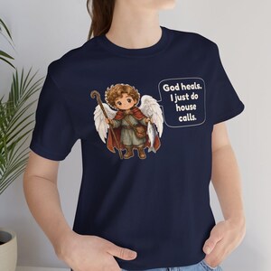 May include: A navy blue t-shirt featuring a graphic of a winged angel figure holding a wooden staff and a fish. The angel wears a red cloak and brown boots. A speech bubble next to the figure contains the text: God heals. I just do house calls.