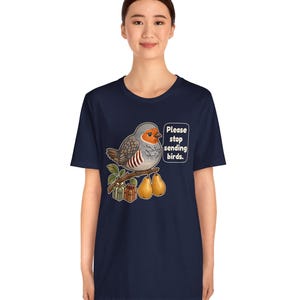 K&ouml;nnte beinhalten: Ein dunkelblaues Kurzarm-T-Shirt mit einer Grafik eines grauen und orangefarbenen Vogels, der auf einem Ast mit zwei Birnen und zwei kleinen Geschenkkartons sitzt. Eine Sprechblase neben dem Vogel enth&auml;lt den Text, Please stop sending birds.