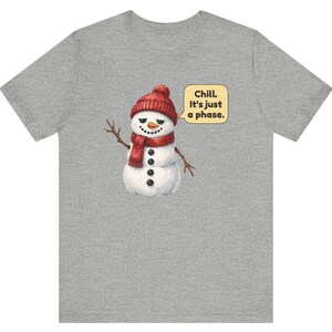 Pode incluir: Uma t-shirt cinzenta mesclada com um gr&aacute;fico de um boneco de neve a usar um gorro e um cachecol de malha vermelha. O boneco de neve tem um nariz de cenoura, bra&ccedil;os de pau e bot&otilde;es pretos. Um bal&atilde;o de fala cont&eacute;m o texto, Chill. It's just a phase.