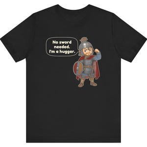 May include: A black t-shirt featuring a cartoon illustration of a warrior in chainmail and a red cape, wearing a helmet with a small animal on top. The warrior holds a round shield and has a speech bubble that says, No sword needed. I'm a hugger.