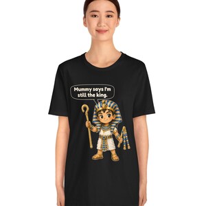 Może przedstawiać: Czarny t-shirt z grafiką przedstawiającą rysunkowego faraona trzymającego laskę i bicz. Nad postacią znajduje się dymek z tekstem: "Mummy says I'm still the king." Faraon ma na sobie niebiesko-złote nakrycie głowy w paski oraz białą tunikę.