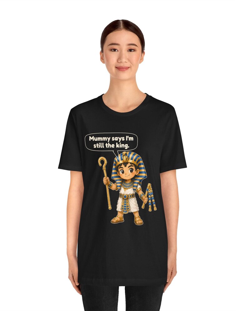Może przedstawiać: Czarny t-shirt z grafiką przedstawiającą rysunkowego faraona trzymającego laskę i bicz. Nad postacią znajduje się dymek z tekstem: "Mummy says I'm still the king." Faraon ma na sobie niebiesko-złote nakrycie głowy w paski oraz białą tunikę.