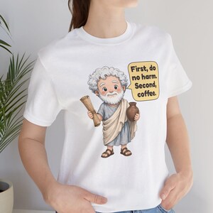 May include: A white t-shirt featuring a cartoon illustration of an elderly man with a white beard wearing a beige toga and brown sandals. He holds a scroll and a brown jug. A yellow speech bubble contains the text: First, do no harm. Second, coffee.