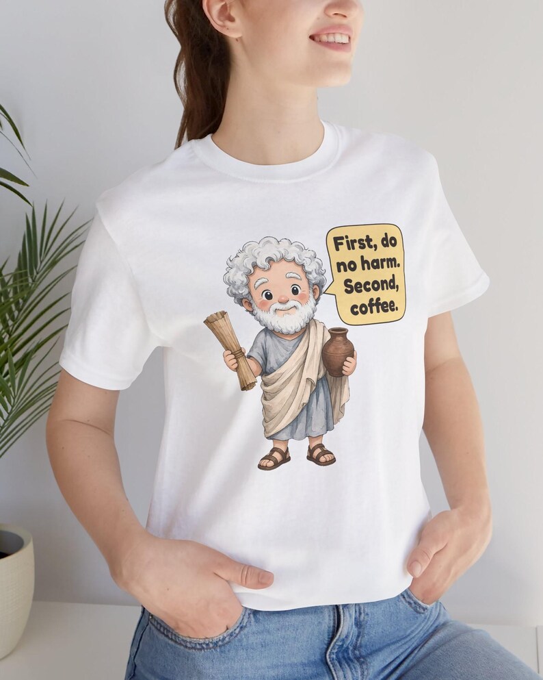 May include: A white t-shirt featuring a cartoon illustration of an elderly man with a white beard wearing a beige toga and brown sandals. He holds a scroll and a brown jug. A yellow speech bubble contains the text: First, do no harm. Second, coffee.