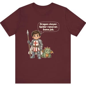 K&ouml;nnte beinhalten: Ein bordeauxrotes Kurzarm-T-Shirt mit einer Grafik eines Cartoon-Ritters in silberner R&uuml;stung, der einen Speer neben einem kleinen gr&uuml;nen Drachen h&auml;lt. Eine Sprechblase &uuml;ber dem Ritter enth&auml;lt den Text: Dragon slayer. Spider remover. Same job.