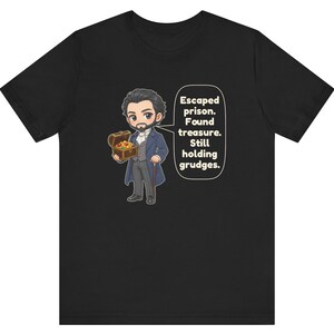 May include: A black t-shirt featuring a cartoon illustration of a bearded man in a blue coat holding a treasure chest. A speech bubble next to the character contains the text: Escaped prison. Found treasure. Still holding grudges.