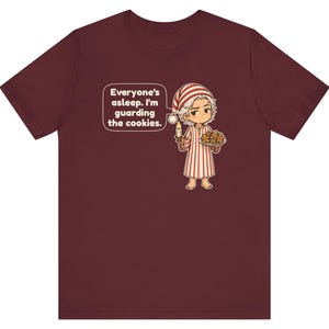 Op de afbeelding: Een bordeauxrood t-shirt met korte mouwen en een stripfiguur in een gestreepte pyjama en slaapmuts, met een kaars en een bord koekjes. Een tekstballon bevat de tekst: Everyone's asleep. I'm guarding the cookies.