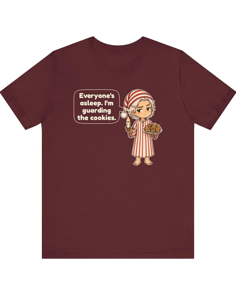 Op de afbeelding: Een bordeauxrood t-shirt met korte mouwen en een stripfiguur in een gestreepte pyjama en slaapmuts, met een kaars en een bord koekjes. Een tekstballon bevat de tekst: Everyone's asleep. I'm guarding the cookies.