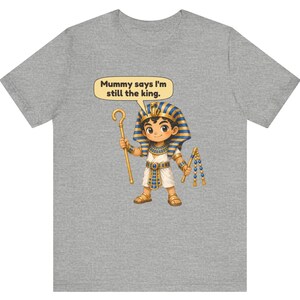 Może przedstawiać: Szary melanżowy t-shirt z rysunkową ilustracją młodego faraona w niebiesko-złotym nakryciu głowy i białej tunice. Postać trzyma złote berło i bicz, z dymkiem zawierającym tekst: Mummy says I'm still the king.