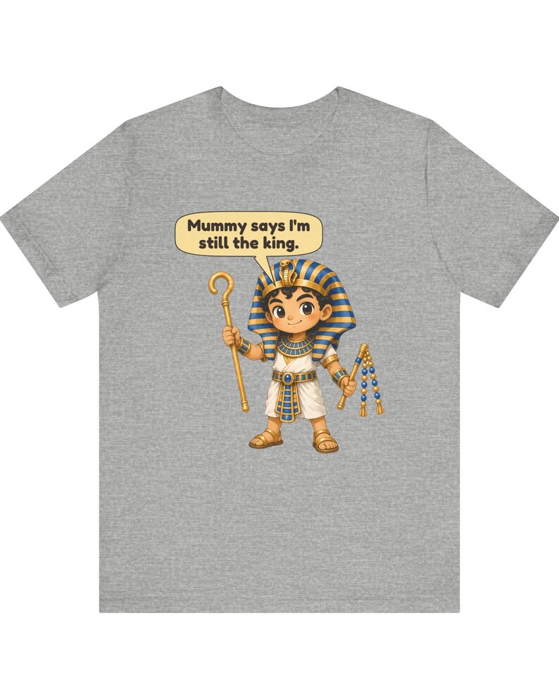 Może przedstawiać: Szary melanżowy t-shirt z rysunkową ilustracją młodego faraona w niebiesko-złotym nakryciu głowy i białej tunice. Postać trzyma złote berło i bicz, z dymkiem zawierającym tekst: Mummy says I'm still the king.
