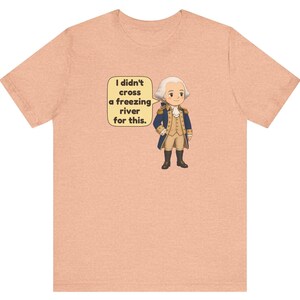 May include: A peach-colored t-shirt featuring a cartoon illustration of a historical figure in a blue and tan military uniform. A speech bubble next to the figure contains the text: I didn't cross a freezing river for this.