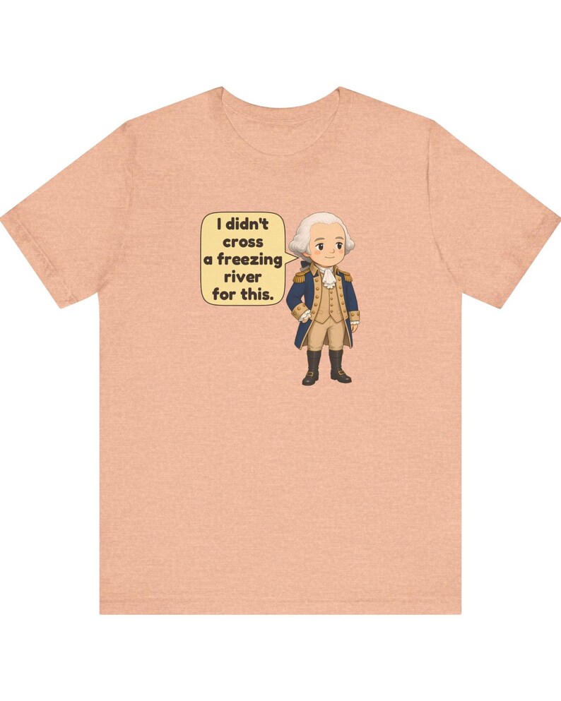 May include: A peach-colored t-shirt featuring a cartoon illustration of a historical figure in a blue and tan military uniform. A speech bubble next to the figure contains the text: I didn't cross a freezing river for this.