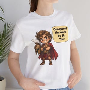 May include: A white t-shirt featuring a cartoon illustration of a warrior in bronze armor and a red cape, holding a plumed helmet. A yellow speech bubble next to the character contains the black text: Conquered the world by 30. You?