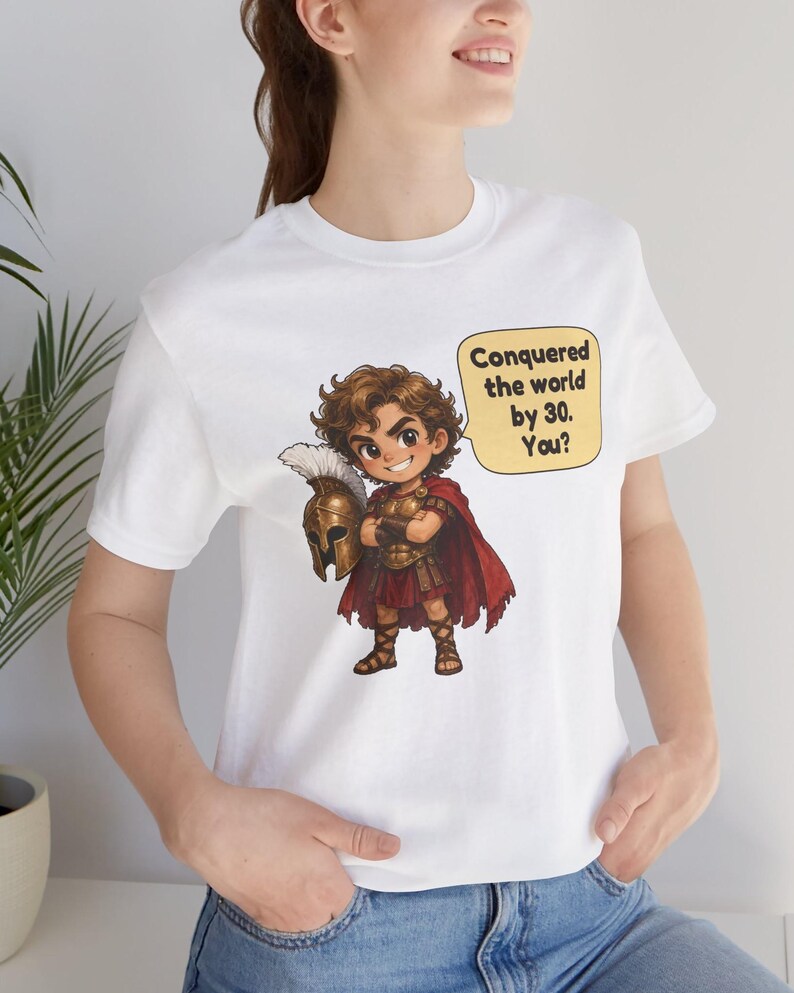 May include: A white t-shirt featuring a cartoon illustration of a warrior in bronze armor and a red cape, holding a plumed helmet. A yellow speech bubble next to the character contains the black text: Conquered the world by 30. You?