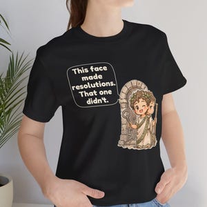 K&ouml;nnte beinhalten: Ein schwarzes T-Shirt mit einer Grafik einer zweigesichtigen r&ouml;mischen Gottheit mit Lorbeerkranz vor einem Steinbogen. Die Figur h&auml;lt einen Schl&uuml;ssel und einen Stab. Eine Sprechblase enth&auml;lt den Text: This face made resolutions. That one didn't.