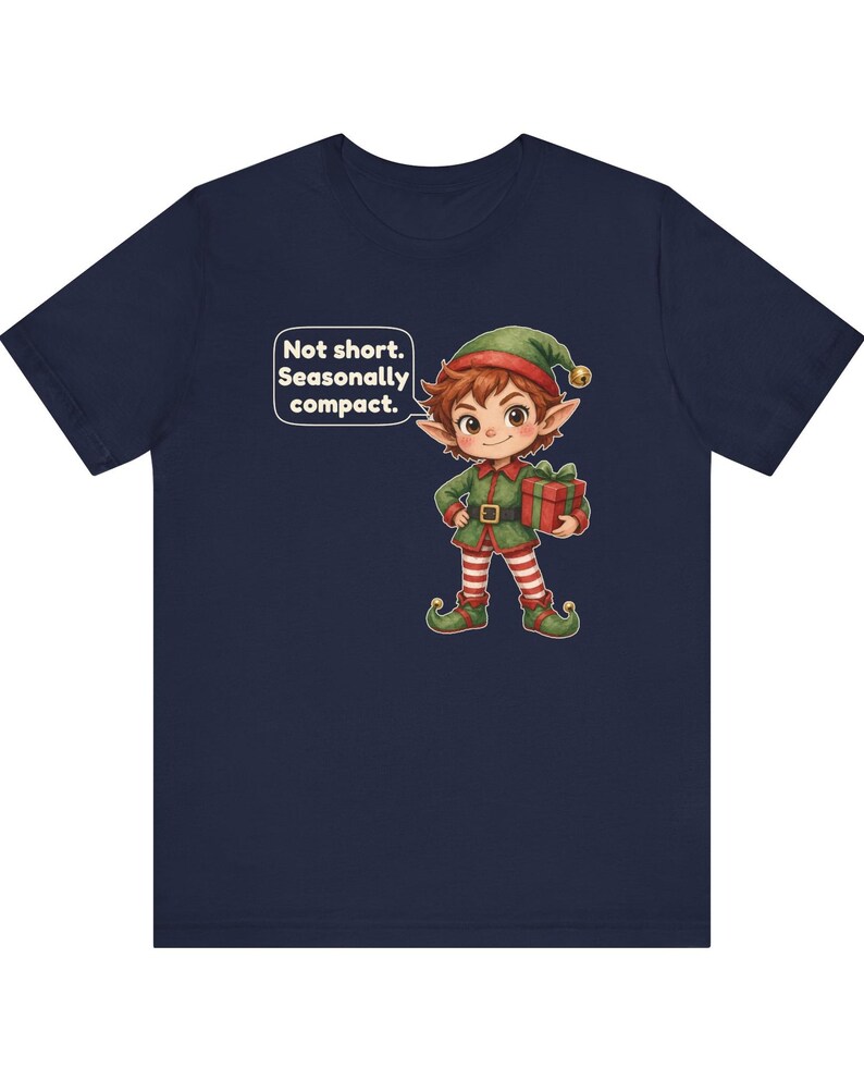 May include: A navy blue t-shirt featuring a cartoon elf wearing a green outfit with red and white striped leggings and a matching hat. The elf holds a red gift box with a green ribbon. A speech bubble contains the text: Not short. Seasonally compact.