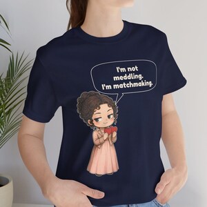 K&ouml;nnte beinhalten: Ein marineblaues T-Shirt mit einer Cartoon-Illustration einer Person mit braunem, lockigem Haar, die eine beige Jacke und ein rosa Kleid tr&auml;gt und zwei rote Herzen h&auml;lt. Eine Sprechblase enth&auml;lt den Text: I'm not meddling. I'm matchmaking.