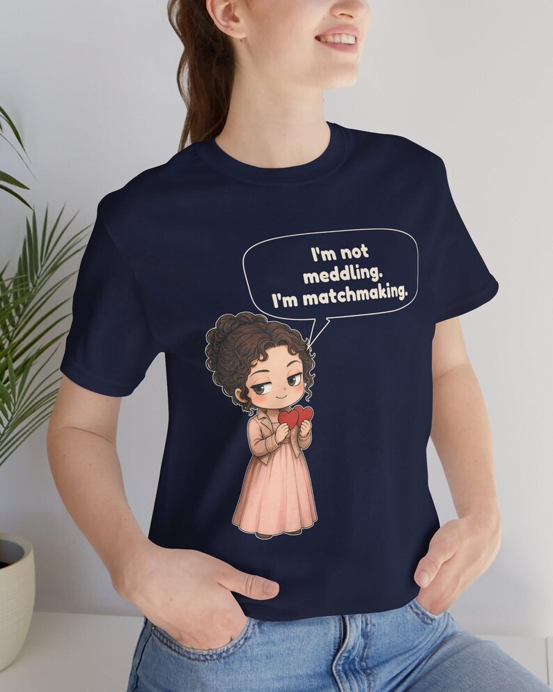 K&ouml;nnte beinhalten: Ein marineblaues T-Shirt mit einer Cartoon-Illustration einer Person mit braunem, lockigem Haar, die eine beige Jacke und ein rosa Kleid tr&auml;gt und zwei rote Herzen h&auml;lt. Eine Sprechblase enth&auml;lt den Text: I'm not meddling. I'm matchmaking.