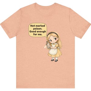 May include: A peach-colored t-shirt featuring a cartoon illustration of a blonde girl in a yellow dress and white apron holding a glass bottle. A speech bubble above her contains the text: Not marked poison. Good enough for me.