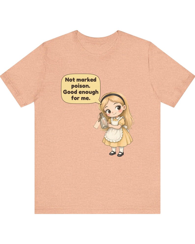 May include: A peach-colored t-shirt featuring a cartoon illustration of a blonde girl in a yellow dress and white apron holding a glass bottle. A speech bubble above her contains the text: Not marked poison. Good enough for me.