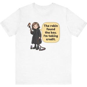 May include: A white t-shirt featuring a cartoon illustration of a person in a black coat holding a large key with a robin perched on it. A speech bubble contains the text: The robin found the key. I'm taking credit. Small purple flowers are visible.