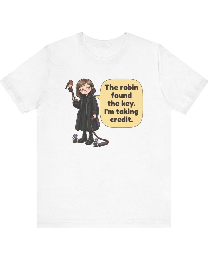 May include: A white t-shirt featuring a cartoon illustration of a person in a black coat holding a large key with a robin perched on it. A speech bubble contains the text: The robin found the key. I'm taking credit. Small purple flowers are visible.