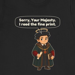 May include: A black t-shirt featuring a cartoon illustration of a person dressed in historical royal attire with a halo, holding a scroll and a book. Above the figure, a speech bubble contains the text: Sorry, Your Majesty. I read the fine print.