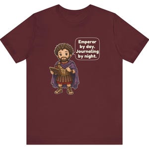 K&ouml;nnte beinhalten: Ein kastanienbraunes T-Shirt mit einer Cartoon-Illustration eines b&auml;rtigen Mannes in r&ouml;mischer R&uuml;stung und einem violetten Umhang, der ein Tagebuch und einen Stift h&auml;lt. Eine Sprechblase neben der Figur enth&auml;lt den Text: Emperor by day. Journaling by night.
