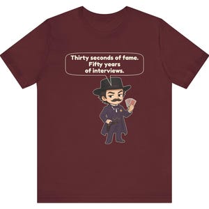 Pu&ograve; includere: Una t-shirt a maniche corte color bordeaux con un'illustrazione a cartone animato di una persona con i baffi, cappello da cowboy e cappotto scuro che tiene in mano delle carte. Un fumetto sopra contiene il testo: Thirty seconds of fame. Fifty years of interviews.