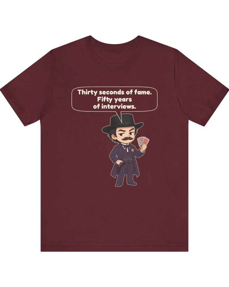 Pu&ograve; includere: Una t-shirt a maniche corte color bordeaux con un'illustrazione a cartone animato di una persona con i baffi, cappello da cowboy e cappotto scuro che tiene in mano delle carte. Un fumetto sopra contiene il testo: Thirty seconds of fame. Fifty years of interviews.