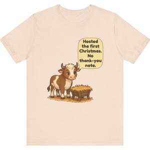 以下が含まれることがあります： 薄いベージュのTシャツ。干し草が入った木製の飼い葉桶の横に立つ、茶色と白の牛のイラストが描かれています。牛の上の吹き出しには「Hosted the first Christmas. No thank-you note.」というテキストがあります。