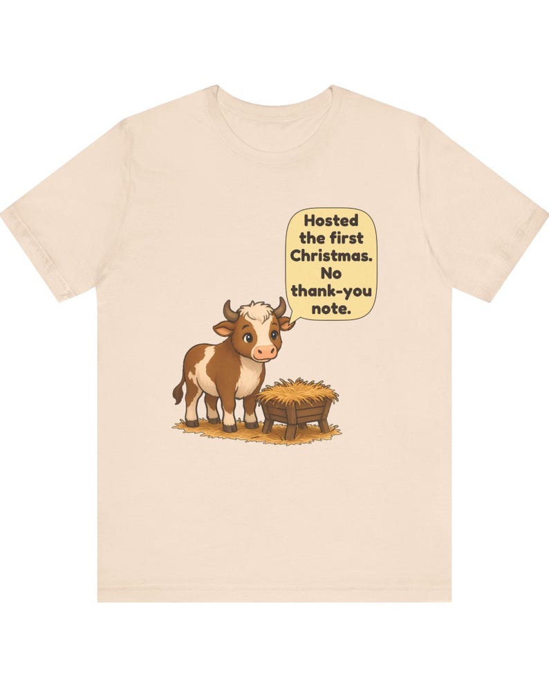 以下が含まれることがあります： 薄いベージュのTシャツ。干し草が入った木製の飼い葉桶の横に立つ、茶色と白の牛のイラストが描かれています。牛の上の吹き出しには「Hosted the first Christmas. No thank-you note.」というテキストがあります。
