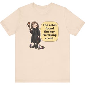 May include: A light beige t-shirt featuring a cartoon illustration of a person in a black coat holding a large key with a robin perched on it. A speech bubble contains the text: The robin found the key. I'm taking credit.