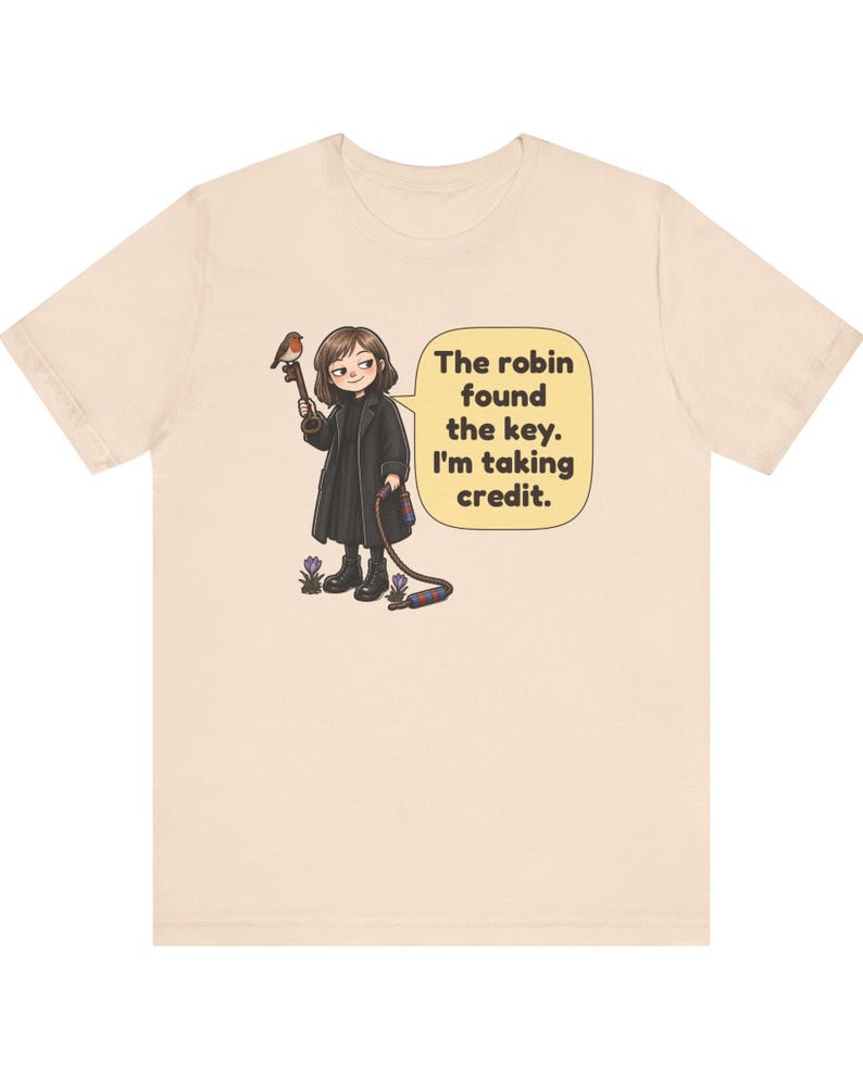 May include: A light beige t-shirt featuring a cartoon illustration of a person in a black coat holding a large key with a robin perched on it. A speech bubble contains the text: The robin found the key. I'm taking credit.