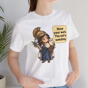 Puede incluir: Una camiseta blanca con el gr&aacute;fico de un personaje con casco dorado y t&uacute;nica azul, sosteniendo una rama de olivo y un libro. Un b&uacute;ho se posa sobre el hombro junto a un bocadillo amarillo con el texto: Show your work. The owl is watching.