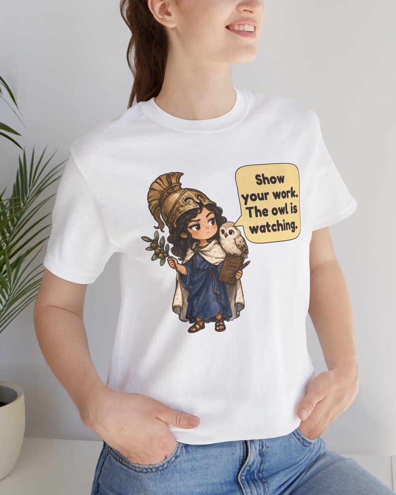Puede incluir: Una camiseta blanca con el gr&aacute;fico de un personaje con casco dorado y t&uacute;nica azul, sosteniendo una rama de olivo y un libro. Un b&uacute;ho se posa sobre el hombro junto a un bocadillo amarillo con el texto: Show your work. The owl is watching.