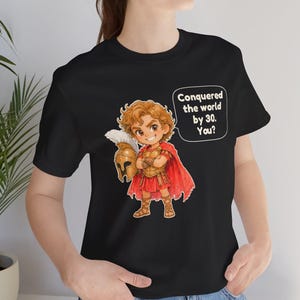 May include: A black t-shirt featuring a cartoon illustration of a historical figure in golden armor and a red cape, holding a helmet. A speech bubble next to the character contains the text: Conquered the world by 30. You?