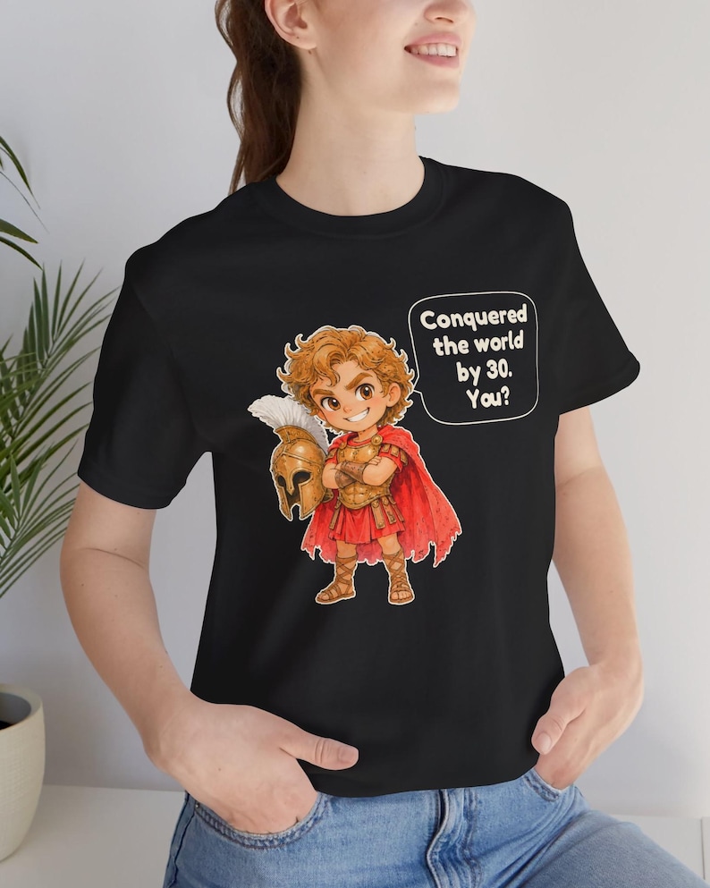May include: A black t-shirt featuring a cartoon illustration of a historical figure in golden armor and a red cape, holding a helmet. A speech bubble next to the character contains the text: Conquered the world by 30. You?