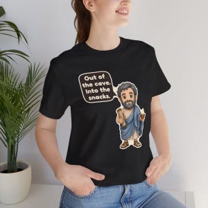 Peut inclure: Un t-shirt noir &agrave; manches courtes avec une illustration de dessin anim&eacute; repr&eacute;sentant un personnage barbu en toge bleue tenant un parchemin et un petit bol. Une bulle contient le texte : Out of the cave. Into the snacks.
