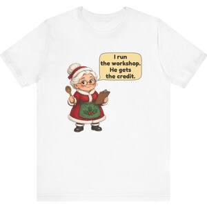 K&ouml;nnte beinhalten: Ein wei&szlig;es T-Shirt mit einer Grafik einer Figur mit wei&szlig;em Dutt, rotem Mantel mit wei&szlig;em Pelzbesatz, gr&uuml;ner Sch&uuml;rze mit Stechpalme und Brille. Die Figur h&auml;lt einen Holzl&ouml;ffel und ein Klemmbrett mit dem Text: I run the workshop. He gets the credit.