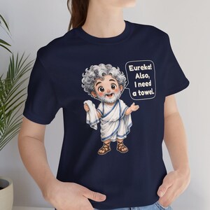 May include: A navy blue t-shirt featuring a cartoon illustration of a bearded man in a white toga and brown sandals. The character holds a white towel and has a speech bubble that reads, "Eureka! Also, I need a towel." The design is centered on the chest.