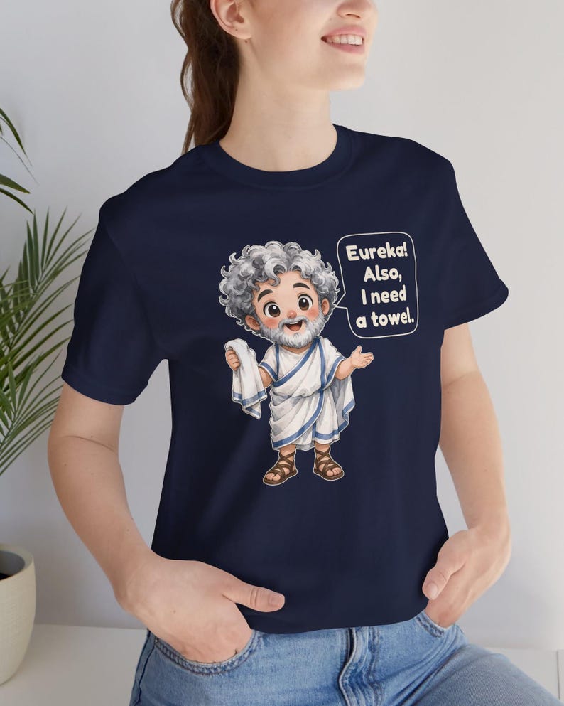 May include: A navy blue t-shirt featuring a cartoon illustration of a bearded man in a white toga and brown sandals. The character holds a white towel and has a speech bubble that reads, "Eureka! Also, I need a towel." The design is centered on the chest.