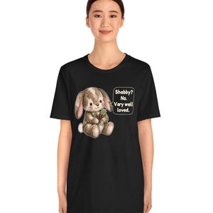May include: A black t-shirt featuring a graphic of a brown stuffed bunny holding a sprig of holly. Next to the bunny is a speech bubble with the text: Shabby? No. Very well loved. The design is centered on the chest of the garment.