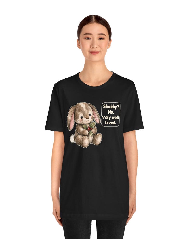 May include: A black t-shirt featuring a graphic of a brown stuffed bunny holding a sprig of holly. Next to the bunny is a speech bubble with the text: Shabby? No. Very well loved. The design is centered on the chest of the garment.